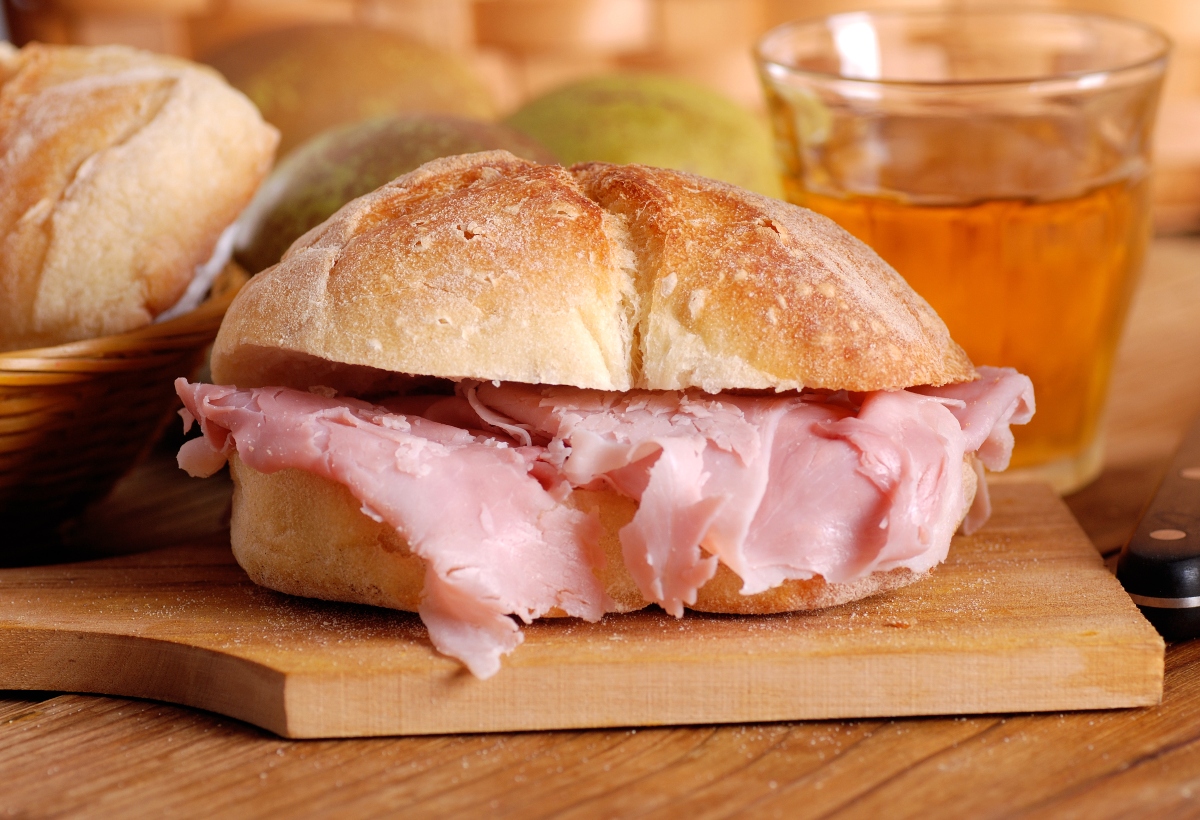 su tagliere di legno primo piano di un panino con prosciutto cotto una delle più famose ricette con prosciutto cotto, dietro un bicchiere di succo di mele e dietro cestino con altri panini