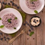 Risotto ai mirtilli servito su piatti bianchi con decorazione di foglie di menta, affiancato da ciotoline con riso, mirtilli freschi e crema al formaggio, su tavolo rustico in legno.