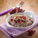 Risotto al melograno: primo piatto semplice e veloce con video-ricetta 1 Piatto di risotto al melograno servito in una ciotola bianca, decorato con chicchi rossi freschi e finocchietto, adagiato su un tovagliolo lilla con forchetta e frutto di melograno a lato.