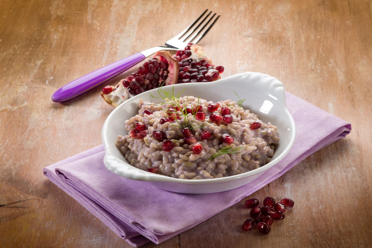 Piatto di risotto al melograno servito in una ciotola bianca, decorato con chicchi rossi freschi e finocchietto, adagiato su un tovagliolo lilla con forchetta e frutto di melograno a lato.