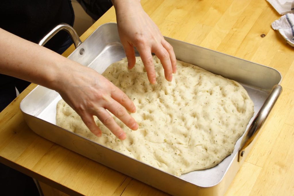 In primo piano delle mani che stendono l’impasto della schiacciata in una teglia rettangolare. La pasta, già lievitata e aromatizzata con erbe, viene pressata con le dita per creare la tipica superficie irregolare prima di essere infornata.