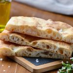Tre fette di schiacciata sovrapposte su un tagliere, con crosta dorata e interno soffice e ben alveolato, accompagnate da sale grosso e olio extravergine di oliva in secondo piano.