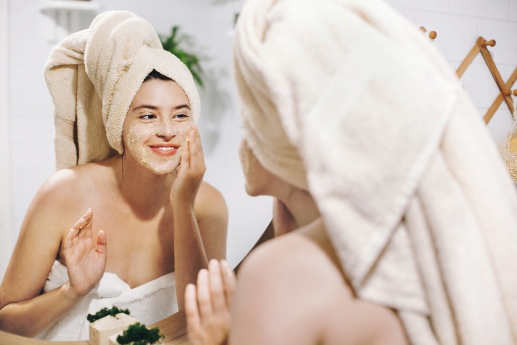 donna allo specchio con asciugamano nei capelli e attorno al corpo. e' sorridente e sta applicando uno scrub viso