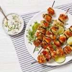 Souvlaki di maiale: la ricetta dello spiedino greco 1 Su una tovaglietta a righe c'รจ un piatto bianco con souvlaki grigliati, spicchi di lime ed erbe aromatiche, serviti accanto a una ciotola di salsa allo yogurt.
