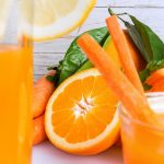 primo piano di un bicchiere di succo ACE con una fette di limone sul bicchiere e sulla destra gli ingredienti per fare un succo pieno di vitamine: arancia, carote e erbe aromatiche