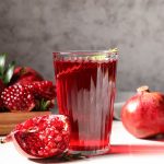 Un bicchiere di succo di melograno dal colore rosso intenso è posizionato al centro, circondato da frutti interi e tagliati che mostrano i chicchi lucidi.