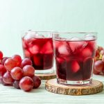 Due bicchieri di succo d’uva rossa con ghiaccio poggiati su sottobicchieri in legno, affiancati da grappoli di uva fresca su un tavolo chiaro.