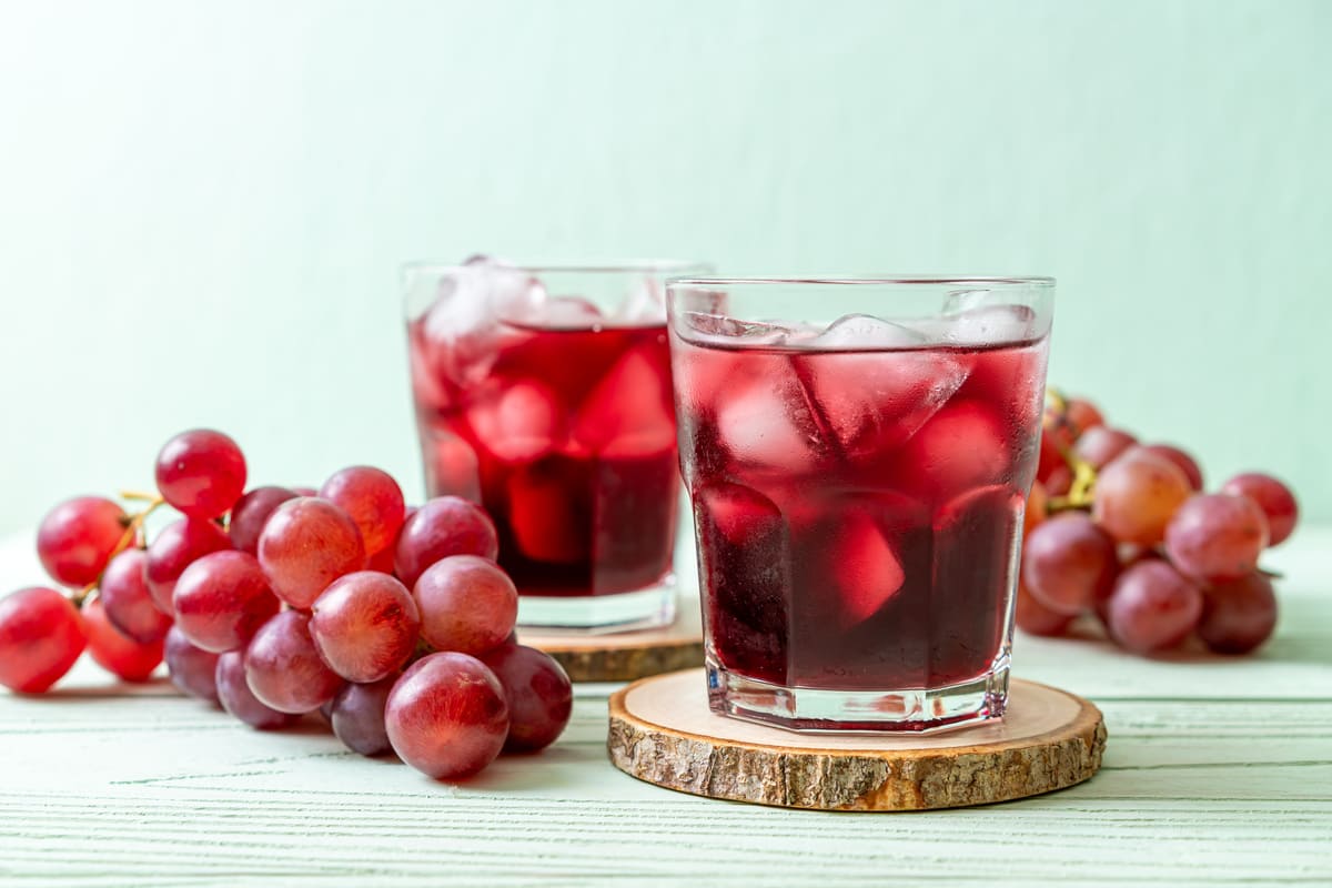 Due bicchieri di succo d’uva rossa con ghiaccio poggiati su sottobicchieri in legno, affiancati da grappoli di uva fresca su un tavolo chiaro.