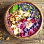 su tavolo di legno: ciotola di legno con un acai bowl colorata con yogurt greco, semi di chia, bacche di acai, mandorle, frutti di bosco e banane. Accanto cucchiaino con frutta secca