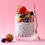 su sfondo rosa, in un bicchiere trasparente un chia pudding con semi di chia, frutta tagliata a pezzi, mirtilli, fragole,kiwi