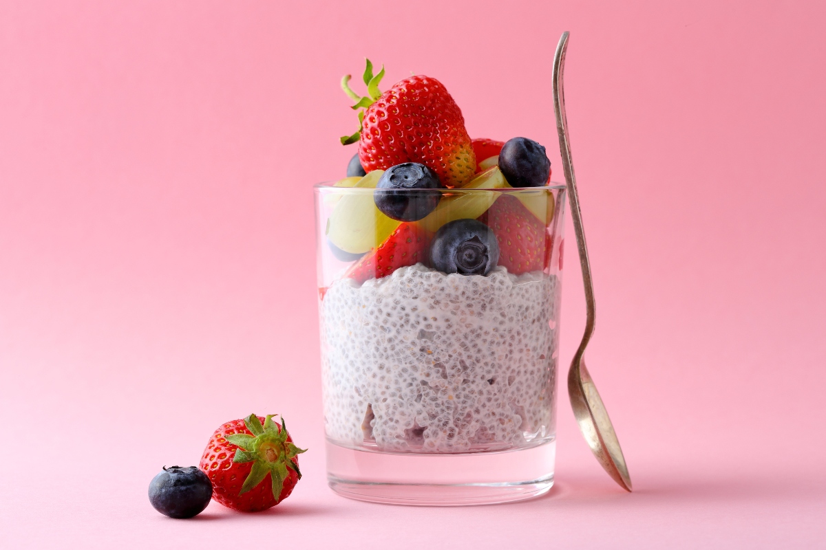 su sfondo rosa, in un bicchiere trasparente un chia pudding con semi di chia, frutta tagliata a pezzi, mirtilli, fragole,kiwi