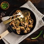 In primo piano una padella con funghi champignon in padella, conditi con erba cipollina fresca e accompagnati da sale rosa in granelli.