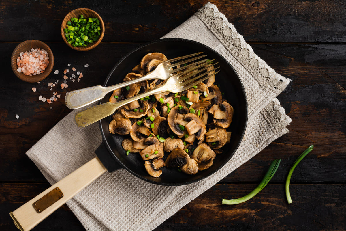In primo piano una padella con funghi champignon in padella, conditi con erba cipollina fresca e accompagnati da sale rosa in granelli.