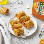 su tavolo con tovaglia bianca spiedini di involtini di zucca con accanto la scatola di pane azzimo integrale di Céréal e attorno una bottiglia di olio e altri spiedini