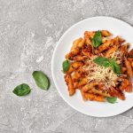 In primo piano un piatto di pasta alla pizzaiola con passata di pomodoro, formaggio grattugiato e foglie di basilico fresco su sfondo grigio.