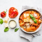 In primo piano un piatto di pasta allo scarpariello con pomodoro, basilico fresco e scaglie di formaggio servito in una ciotola chiara su tovagliolo di lino, con ingredienti freschi come pomodori, peperoncino e formaggio accanto.