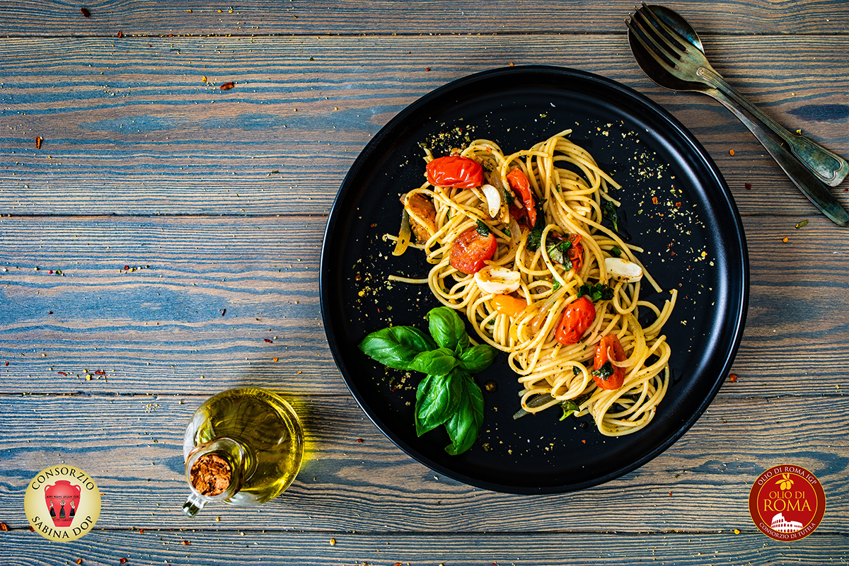 su tavolo di legno azzurro, piatto nero con una delle migliori ricette con l'olio extravergine d'oliva: pasta al pomodoro e basilico sulla sinistra una bottiglia di olio e in basso a destra e sinistra loghi olio Roma IGP e olio Sabina DOP