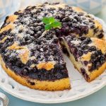 In primo piano c’è una torta ai mirtilli soffice, decorata con zucchero a velo e servita su un piatto bianco.