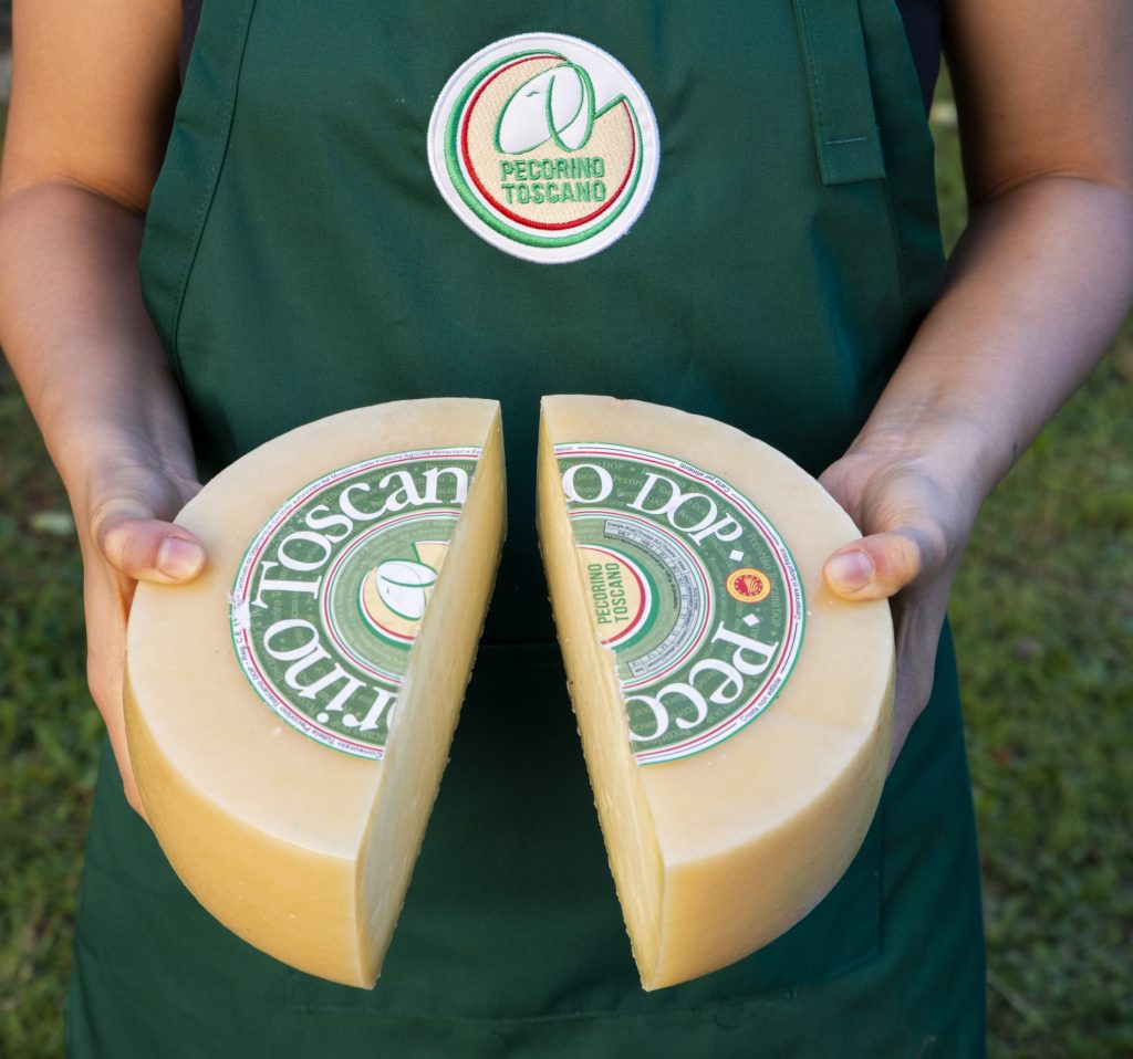Pecorino: proprietà nutrizionali, benefici, ricette e come inserirlo a dieta 2 donna con grembiule verde e logo del pecorino Toscano che tiene in mano un formaggio tagliato in 2