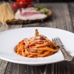 Bucatini all'amatriciana: la ricetta originale facile e veloce 2 Su un tavolo di legno scuro, in un piatto fondo bianco ci sono dei gustosi bucatini all'amatriciana. Sullo sfondo ci sono gli ingredienti principali della ricetta: pasta, guanciale e pomodori.