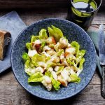 su tavolo di legno insalata di pollo con la crema di Pecorino in una ciotola blu. Sulla sinistra un pezzo di pecorino toscano DOP su tagliere blu e sulla destra tovagliolo blu con forchetta e coltello. Dietro un bicchiere verde di acqua