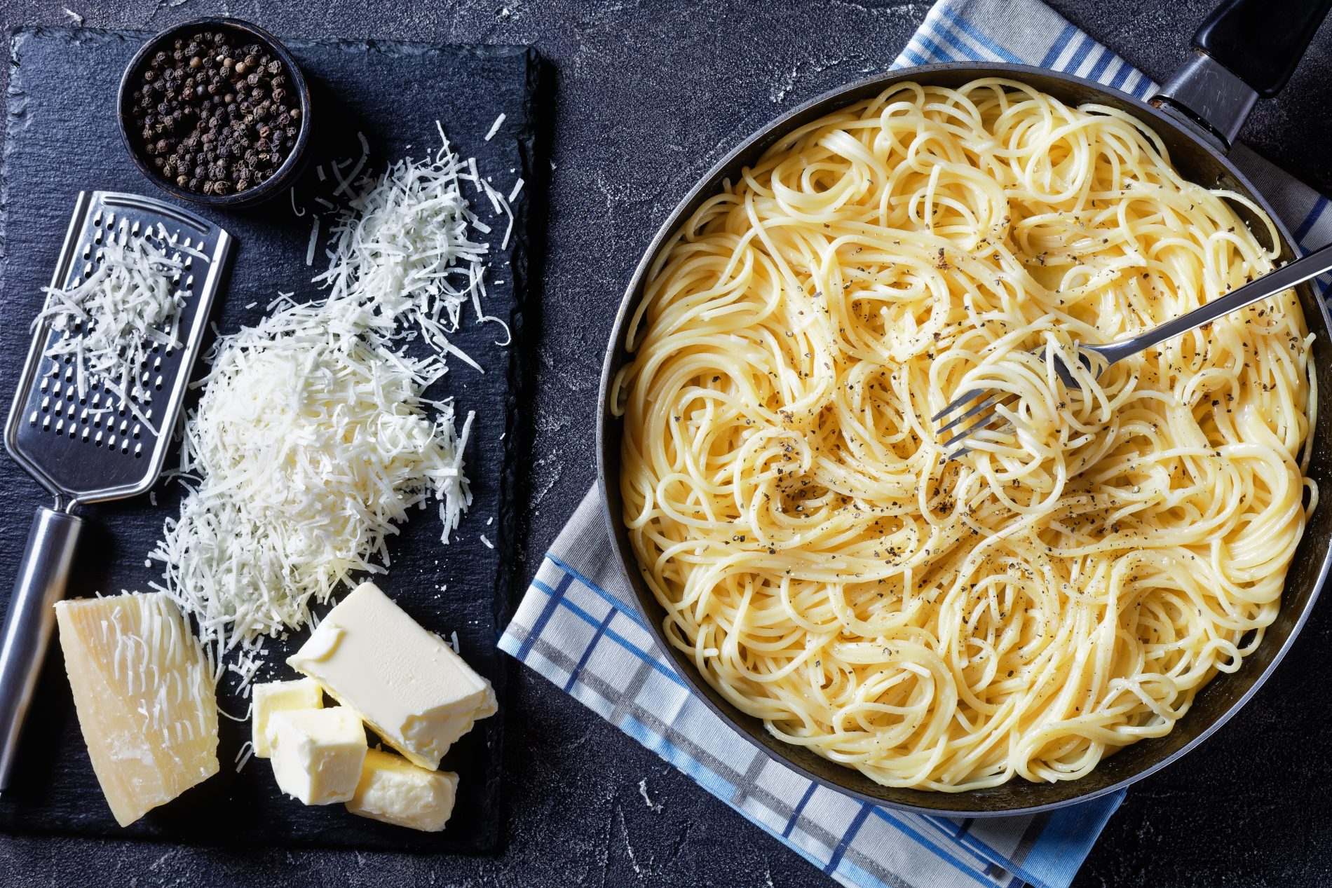 foto con a destra un piatto di spaghetti in una ciotola nera sotto un canovaccio bianco a righe blu mentre sulla sinistra un tagliere nero con pepe nero una grattugia e pecorino grattugiato e pezzi di pecorino