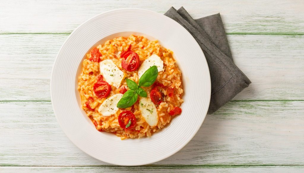 Riso alla pizzaiola: un primo piatto semplice, sano e delizioso 2 su tavolo legno verde chiaro, piatto bianco fondo co riso alla pizzaiola con pomodori, mozzarella e basilico