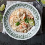In primo piano su un tavolo di legno, c'è un piatto fondo con delle decorazioni verdi, all'interno un cremoso risotto agli agrumi e gamberi con uno spicchio di limone. Sullo sfondo un piattino con degli spicchi di limone a destra, mentre sulla sinistra delle erbette aromatiche.
