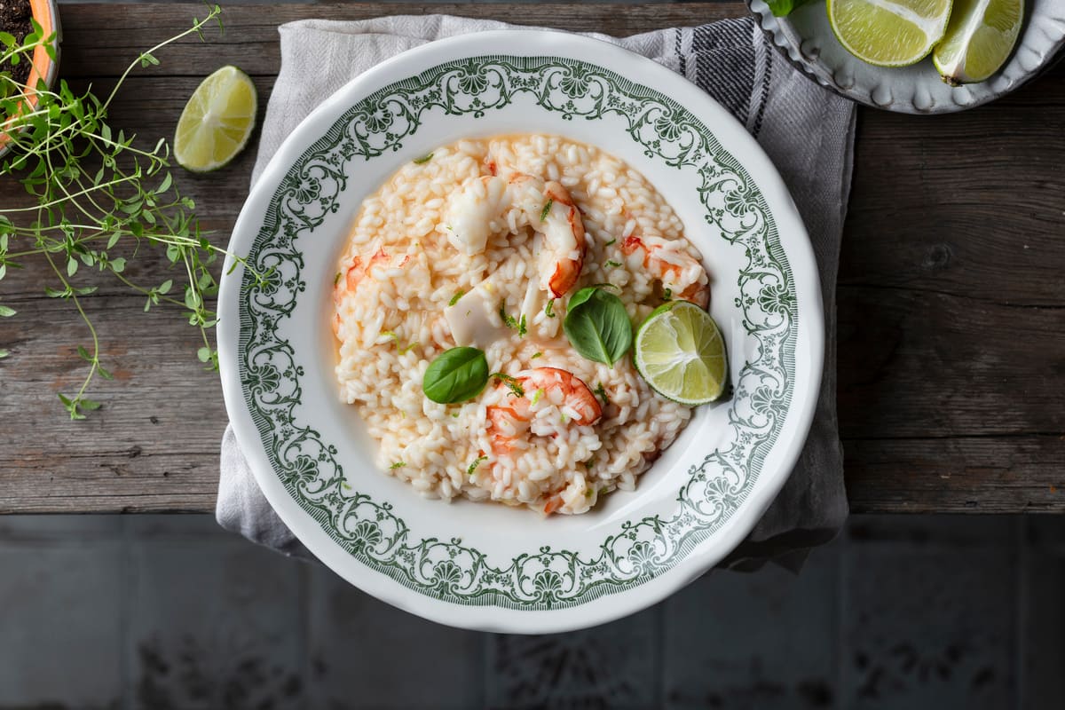 In primo piano su un tavolo di legno, c'è un piatto fondo con delle decorazioni verdi, all'interno un cremoso risotto agli agrumi e gamberi con uno spicchio di limone. Sullo sfondo un piattino con degli spicchi di limone a destra, mentre sulla sinistra delle erbette aromatiche.
