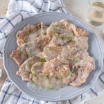 su tavolo, canovaccio e righe bianche e blu sul quale c'è un piatto con scaloppine al vino bianco, con una forchetta sulla destra e un bicchiere di vino bianco e sulla sinistra un cucchiaino con la farina