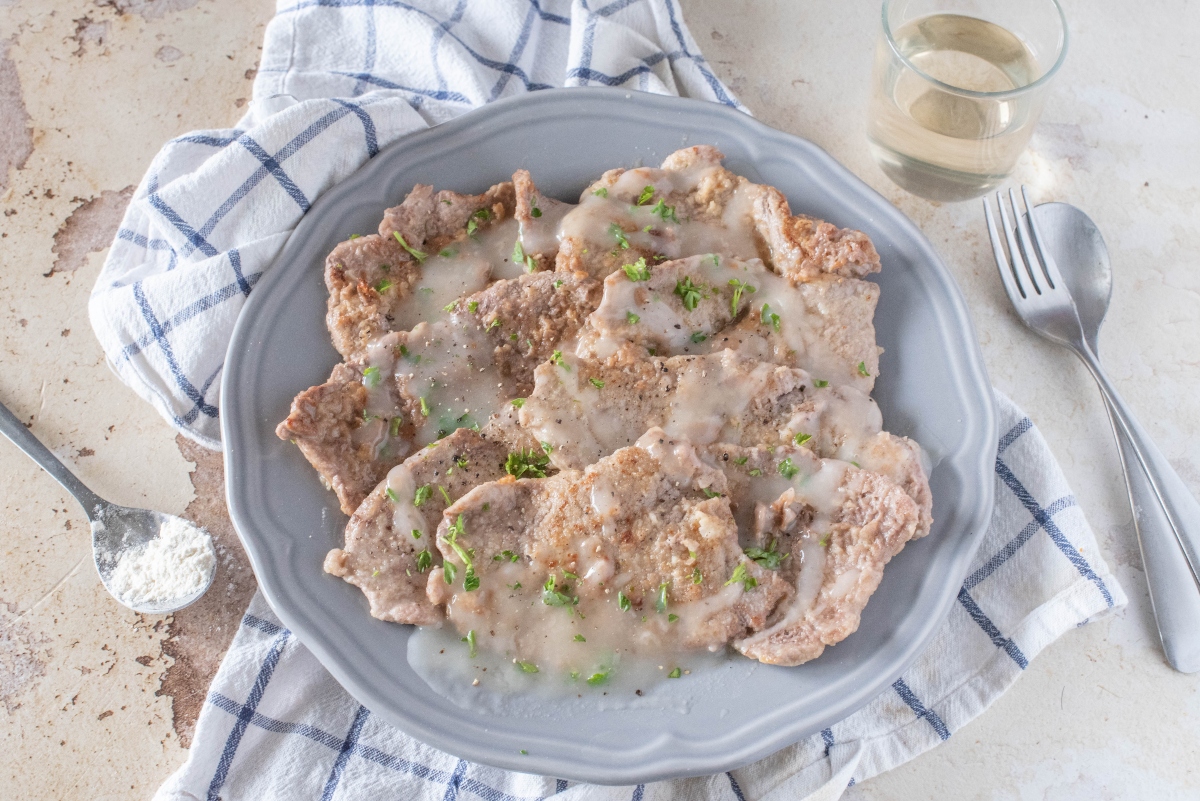 su tavolo, canovaccio e righe bianche e blu sul quale c'è un piatto con scaloppine al vino bianco, con una forchetta sulla destra e un bicchiere di vino bianco e sulla sinistra un cucchiaino con la farina