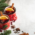 Vin brulé fatto in casa: bevanda calda e speziata con videoricetta 1 In primo piano, sulla sinistra ci sono delle tazze rosse piene di vin brulé con arancia, cannella e anice stellato su uno sfondo chiaro, decorato con degli aghi di pino.