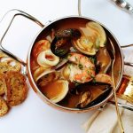 Brodetto di pesce: zuppa dell’Adriatico con pesce misto e varianti regionali 1 In primo piano su un tavolo bianco, in un tegame c'è un brodetto di pesce con gamberi, calamari e cozze. Sulla sinistra ci sono alcune fette di pane tostato mentre sulla destra un tovagliolo color crema.