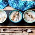 su tavolo di legno. tagliere di ardesia con 3 ciotole con risolatte; la prima con frutta secca, la seconda con una bacca di vaniglia e la terza con cucchiaio. Attorno canovaccio azzurro con righe bianche