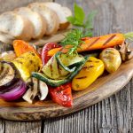 Verdure in friggitrice ad aria: un contorno vegetariano sano e gustoso 1 su tavolo di legno, tagliere di legno con verdure in friggitrice ad aria. Dietro su tagliere più piccolo fette di pane