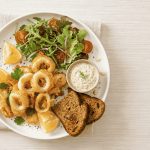Calamari in friggitrice ad aria: una frittura leggera e croccante 1 Piatto con calamari in friggitrice ad aria, insalata mista con pomodorini, salsa allo yogurt, pane tostato e spicchi di limone.