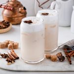 Chai latte: ricetta del tè nero indiano speziato dal gusto avvolgente 1 In primo piano su un tagliere in marmo, due bicchieri in vetro con il chai latte. Intorno ci sono delle spezie come la cannella, chiodi di garofano, una teiera in rame e due piccole caraffe in ceramica bianca.