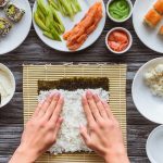 Riso per sushi : come cucinarlo senza errori 1 su tavolo di legno mani di una donna che fa vedere come cucinare il riso sushi con alga le stende con le mani. Dietro ricette di sushi e accanto su piattini salse e riso per sushi