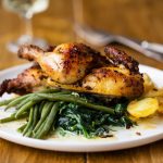 Pollo in friggitrice ad aria: dorato, succoso e pronto in pochi minuti 1 In primo piano su un tavolo di legno chiaro c'รจ un piatto bianco con del pollo con friggitrice ad aria e verdure. Sullo sfondo delle posate e un calice sulla sinistra.