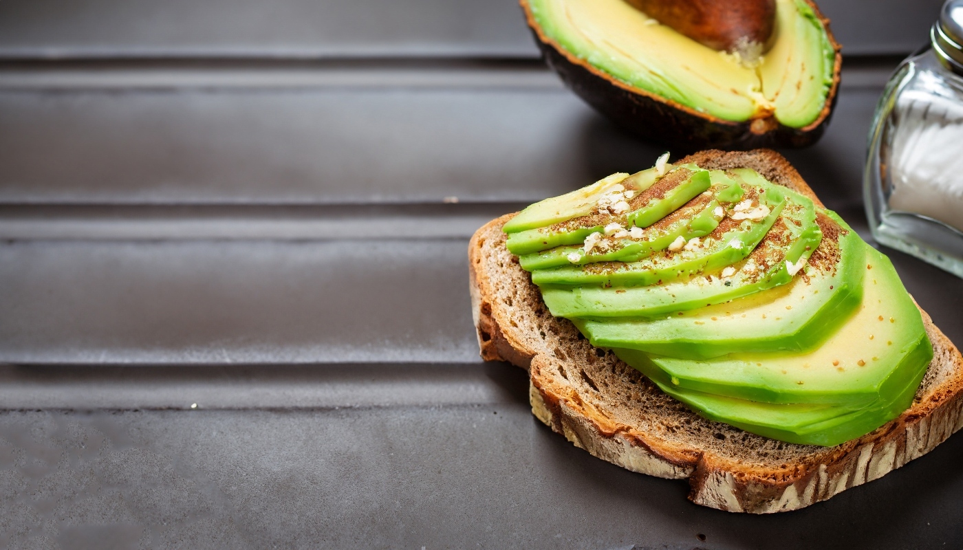 su tavolo fette di pane con avocado tagliato a fettine sottile. Dietro mezzo avocado e la saliera