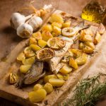 Spigola al cartoccio: ricetta semplice al forno e varianti 1 In primo piano su un tagliere di legno una spigola al cartoccio con patate cotte al forno, fettine di limone. Accanto una treccia di aglio, rosmarino e dell'olio.