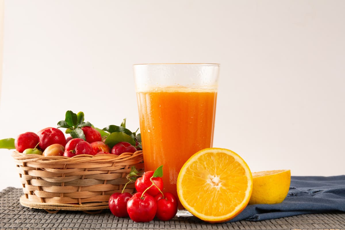 In primo piano, al centro si trova un bicchiere di vetro con del succo di acerola, accanto sulla sinistra c'è una cesta con il frutto dell'acerola. Sulla destra si trova una metà di un'arancia.