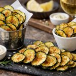 Chips di zucchine al forno: un finger food croccante e sano 1 chips di zucchine servite su vassoio scuro rettangolare e in ciotole, bianca e metallica
