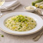 Risotto ai porri: la ricetta per farlo leggero e cremoso 1 tavola apparecchiata con una scodella piena di risotto ai porri, posate e un calice di vino bianco