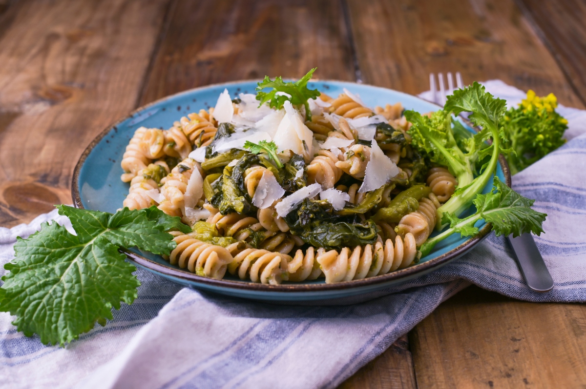 su tavolo di legno, piatto blu con cime di rapa e pasta con sotto un canovaccio bianco a righe blu. Attorno il piatto cine di rapa cruda