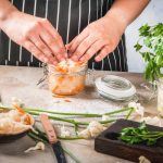 Crauti fatti in casa: ricetta facile del superfood fermentato 1 piano americano di un tavolo di marmo in cucina con le mani di una donna che schiaccia in un contenitore dei crauti fatti in casa. Ha un grembiule nero con righe bianche e attorno altri contenitori e gli ingredienti