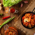 Kimchi: il contorno coreano fermentato fatto in casa 1 su tavolo di legni sotto piatto di paglia con piatto fondo coon il contorno kimchi fatto in casa e attorno ingredienti come cipollato, carote, zenzero, curcuma e peperoni
