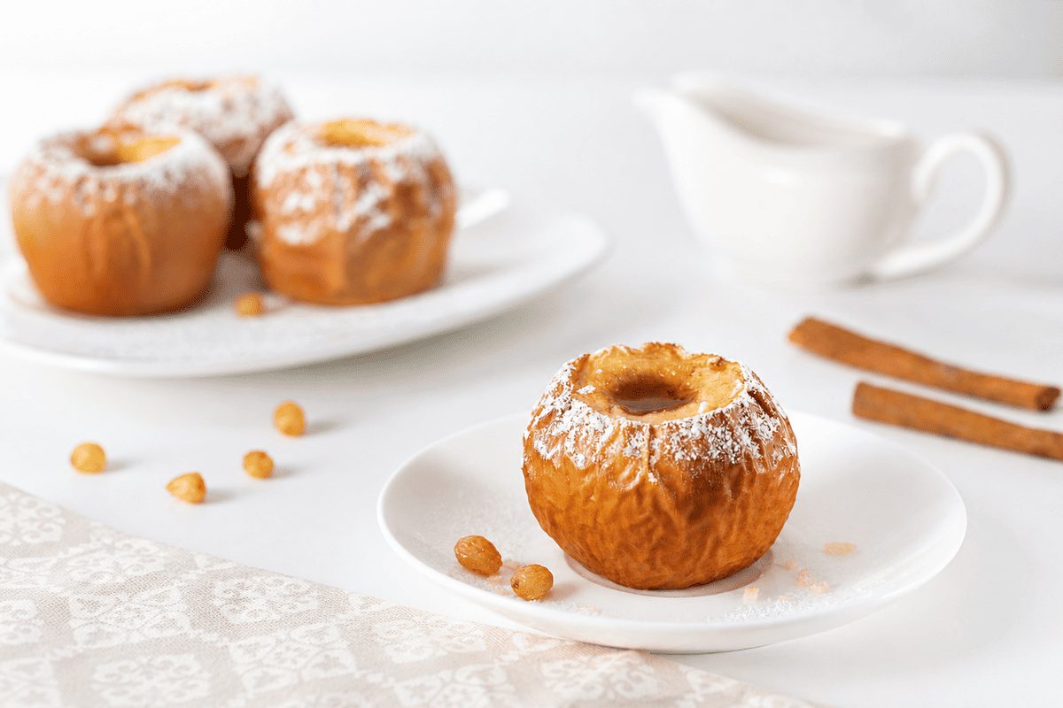 Ricette con le mele: proposte gustose e light 36 In primo piano su un tavolo bianco con una tovaglia chiara ci sono delle mele cotte in friggitrice ad aria. Sullo sfondo due stecche di cannella.