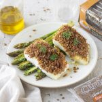 merluzzo in crosta: piatto bianco tondo con due tranci di merluzzo poggiati su alcuni asparagi, una scatola di pane di segale Céréal accanto