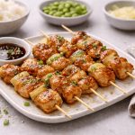 Yakitori: la ricetta degli spiedini di pollo giapponesi 1 In primo piano su un piatto piano quadrato ci sono gli yakitori, degli spiedini di pollo conditi con del porro e la salsa di soia. Sulla destra c'è un tovagliolo beige mentre sullo sfondo ci sono delle ciotoline con alcuni ingredienti della ricetta.